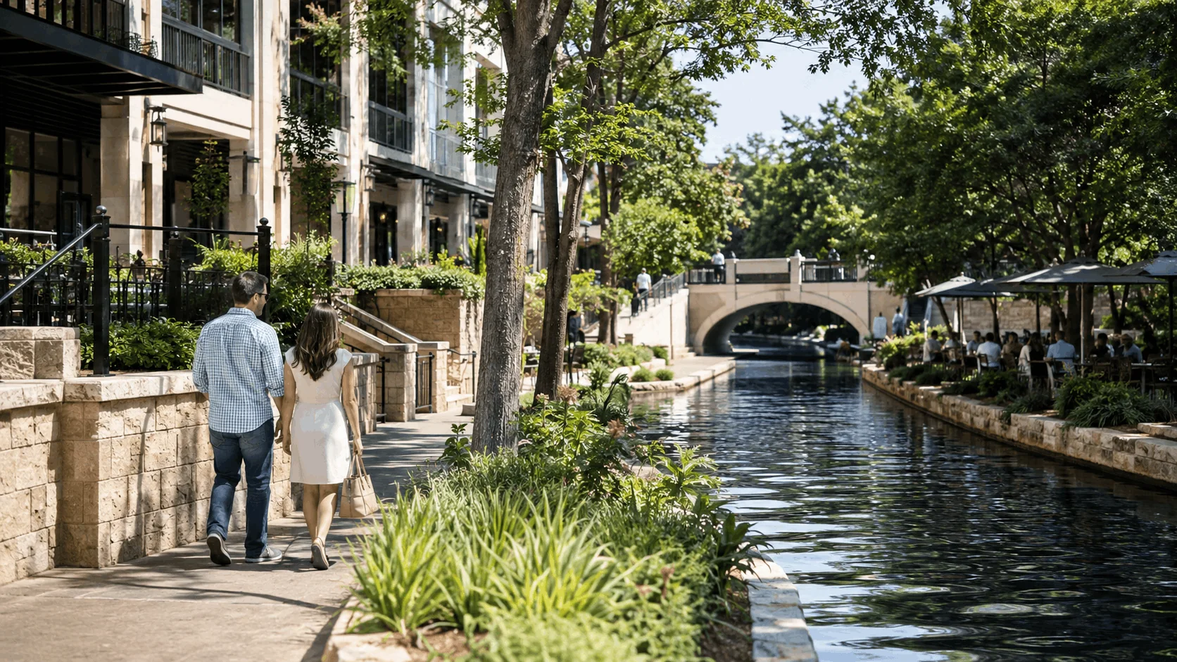 Couple walking along the San Antonio River Walk surrounded by apartments, restaurants, and greenery.