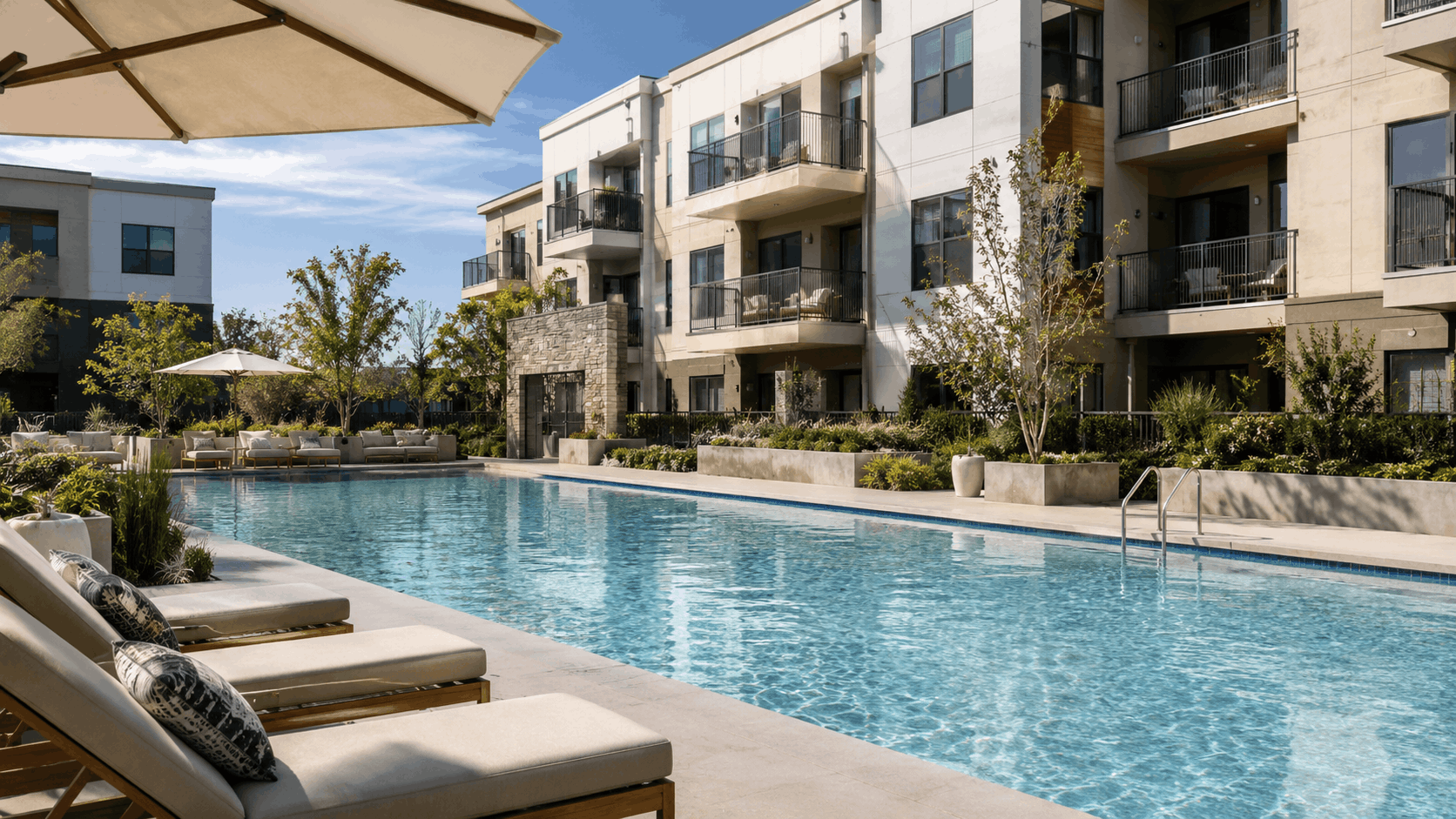 Resort-style apartment pool surrounded by lounge chairs, umbrellas, landscaping, and modern residences.